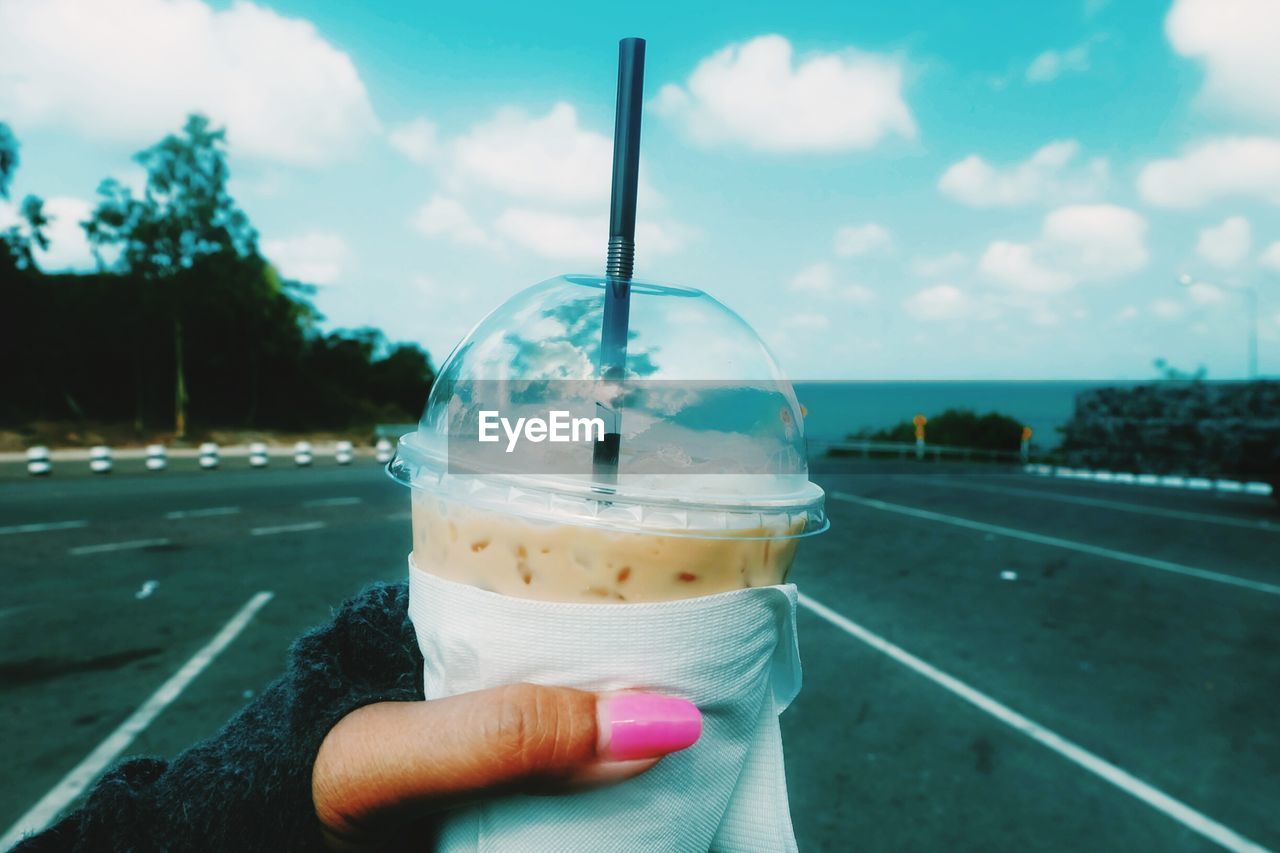 Cropped image of hand holding disposable cup at street against sky