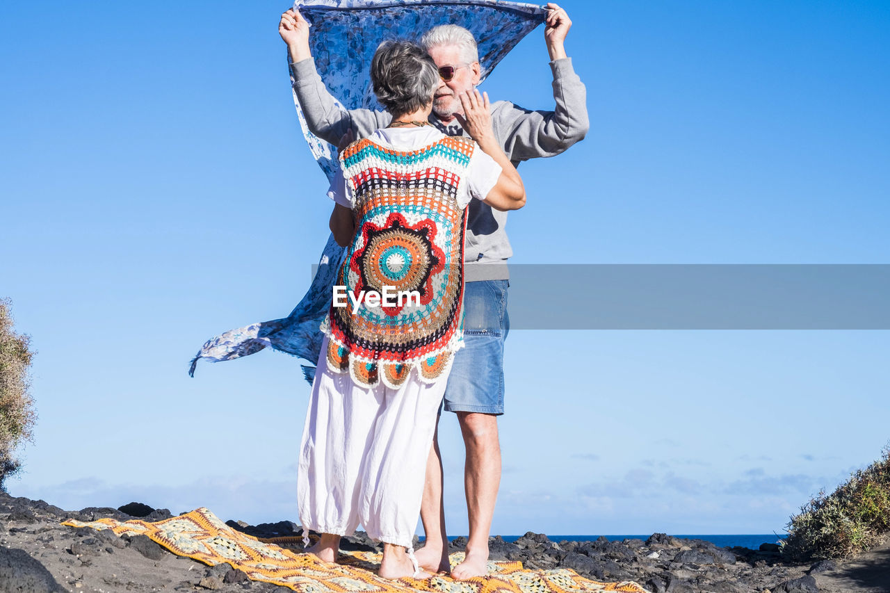 Senior couple standing with sarong at beach against sky