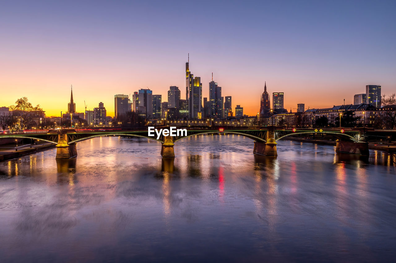 ILLUMINATED BUILDINGS BY RIVER AGAINST SKY IN CITY