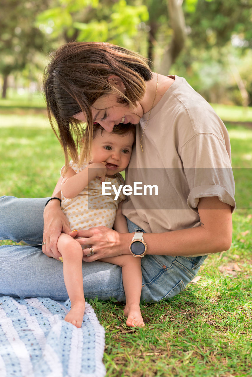 Positive mom sitting on lawn with adorable little daughter while spending pleasant time in park