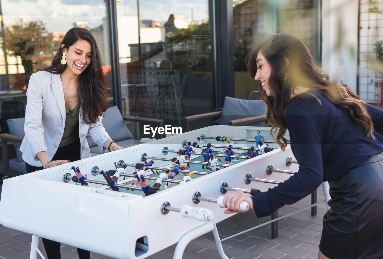 Happy adult hispanic girlfriends in smart casual clothes smiling and playing table football together during party on weekend day on terrace