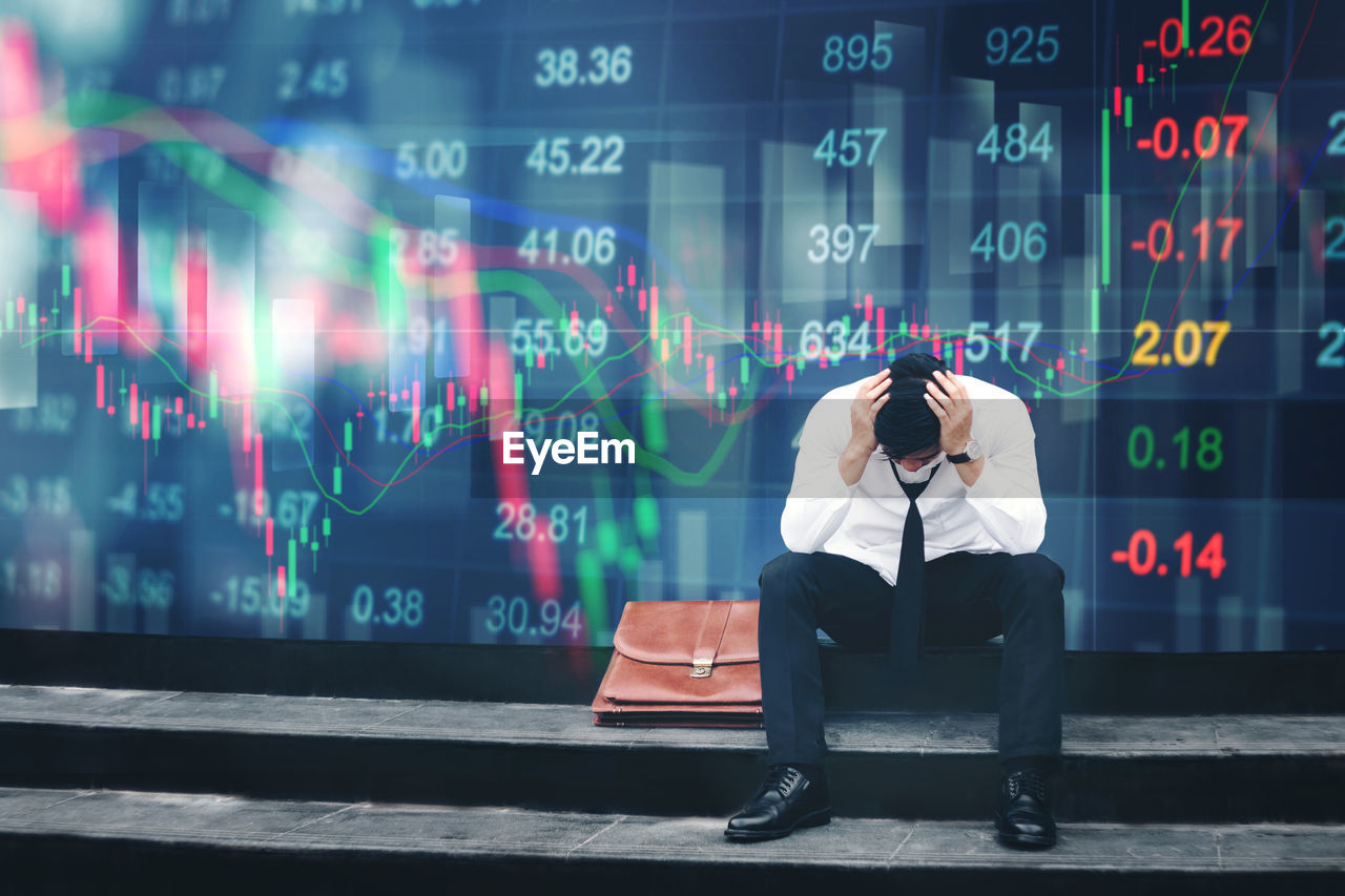 Depressed young businessman with head in hands sitting against stock market data in office