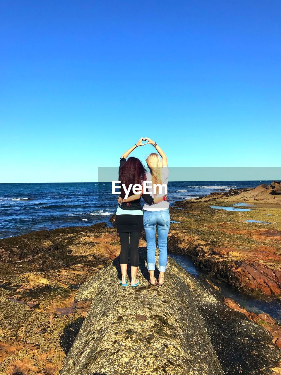 Rear view of women with arms around making heart shape over head while standing on rock at beach against sky