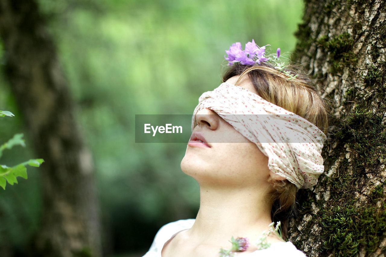 Close-up of woman with blind fold leaning on tree trunk