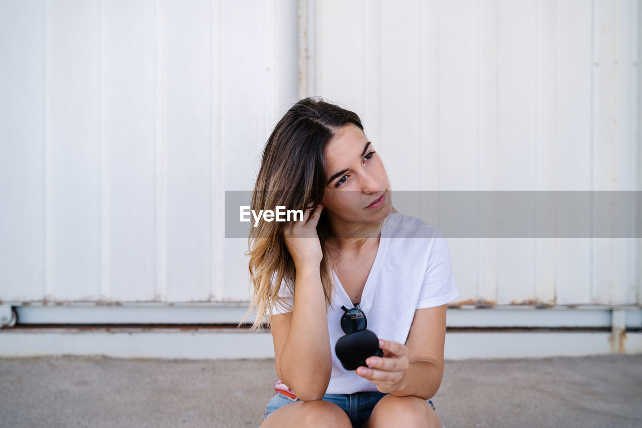 Modern millennial female in casual outfit adjusting wireless earphones while sitting against metal wall on city street