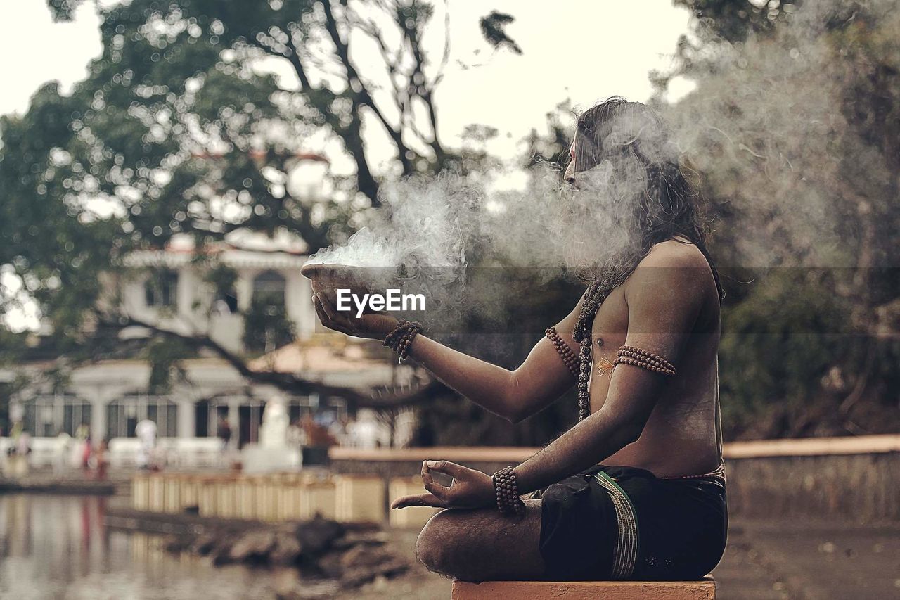 Side view of sadhu sitting with smoke emitting container on stool