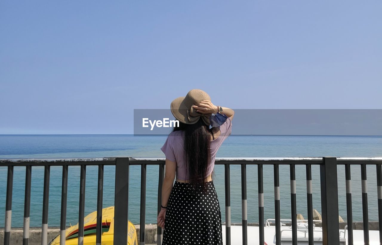 Rear view of woman wearing hat standing by sea against clear sky