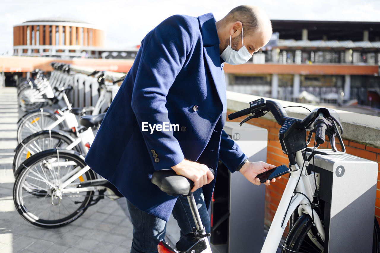 Man wearing protective face mask unlocking bicycle through smart phone on sunny day