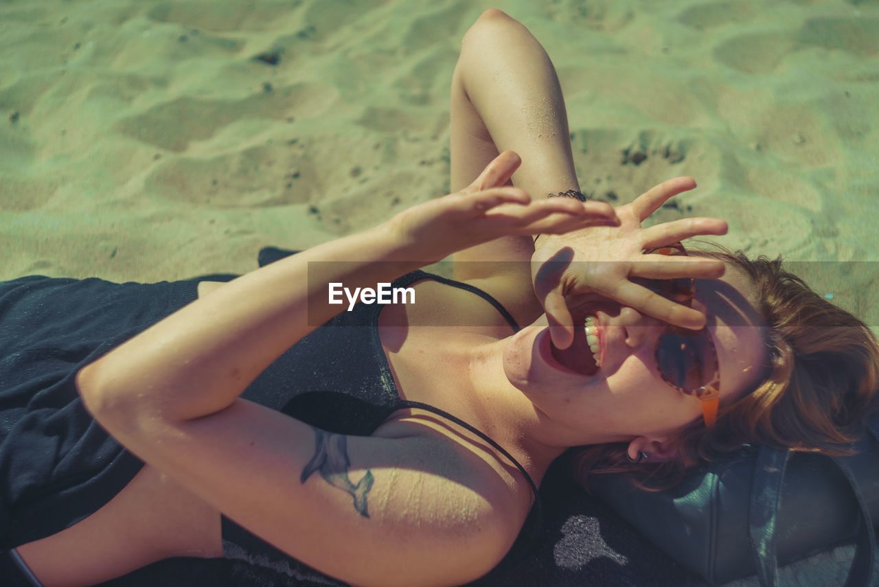 High angle view of cheerful woman lying on sand at beach during sunny day