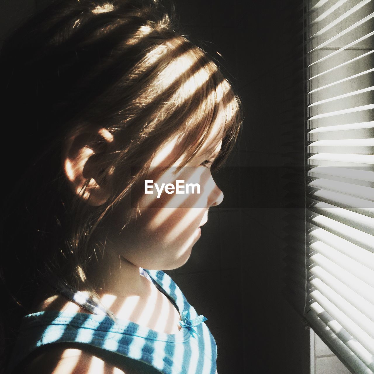 Close-up side view of girl looking through window blinds at home