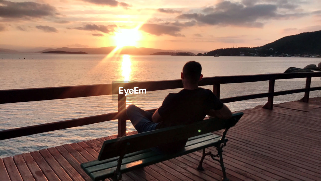 REAR VIEW OF MAN SITTING ON BENCH LOOKING AT SEA