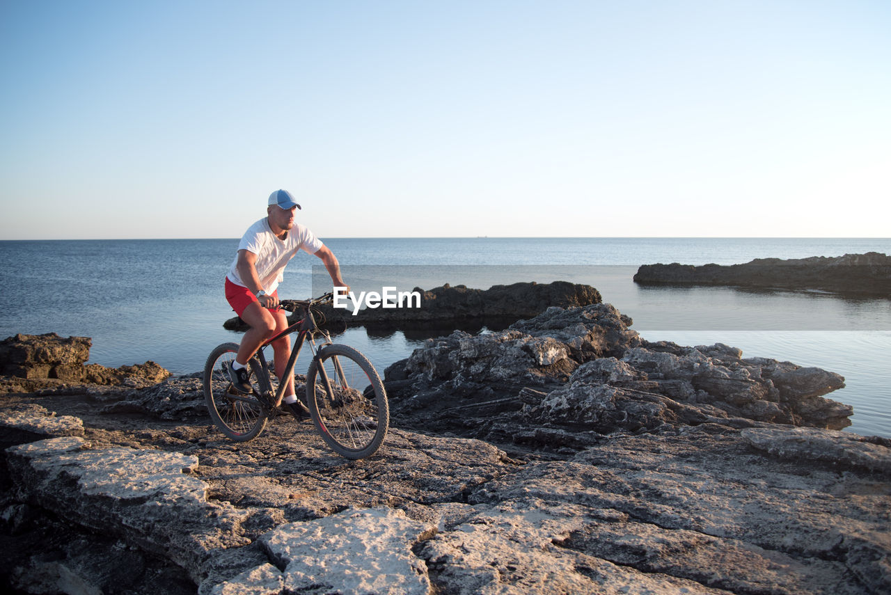 Man riding bicycle on rock by sea against clear sky