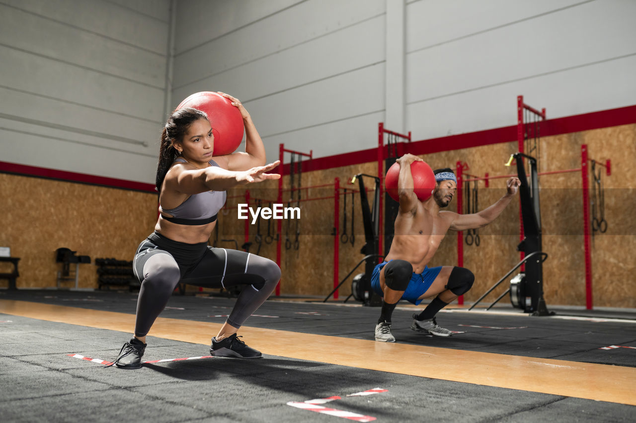 Concentrated sportswoman and sportsman doing squats with heavy medical balls during functional intense training in sports club