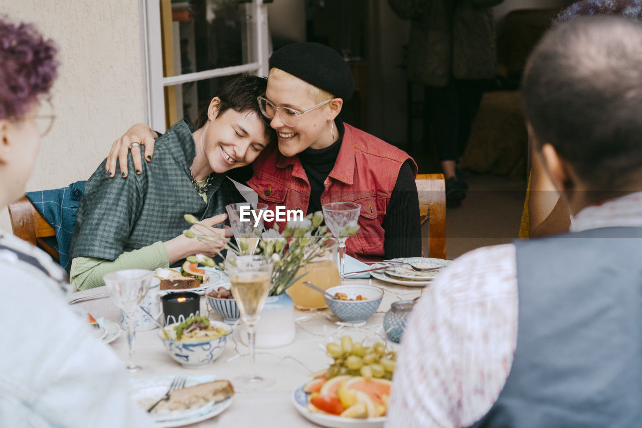 Smiling gay woman sitting with arm around non-binary friend during party in back yard