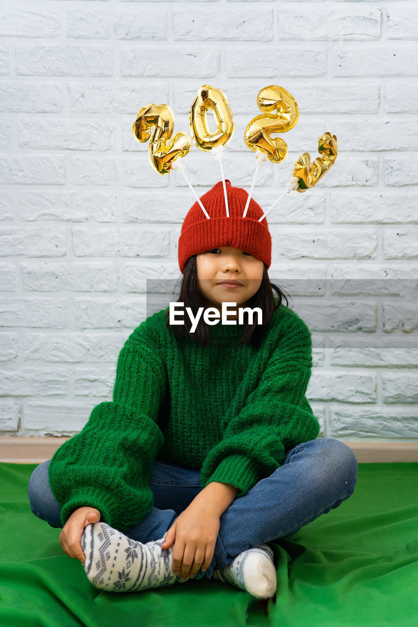 Funny asian girl in green sweater, sits on floor with golden balloons with number 2023 in a red hat