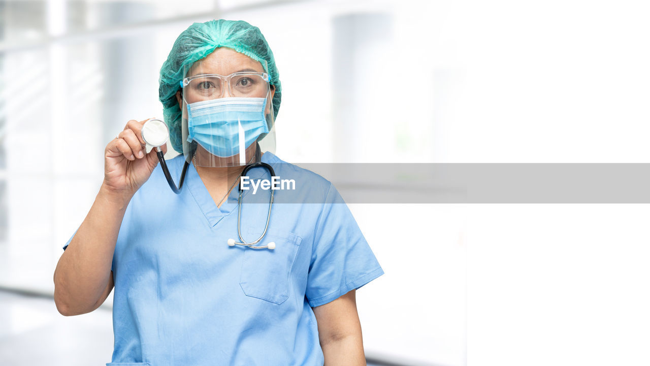 Portrait of doctor wearing mask holding stethoscope standing against wall at hospital