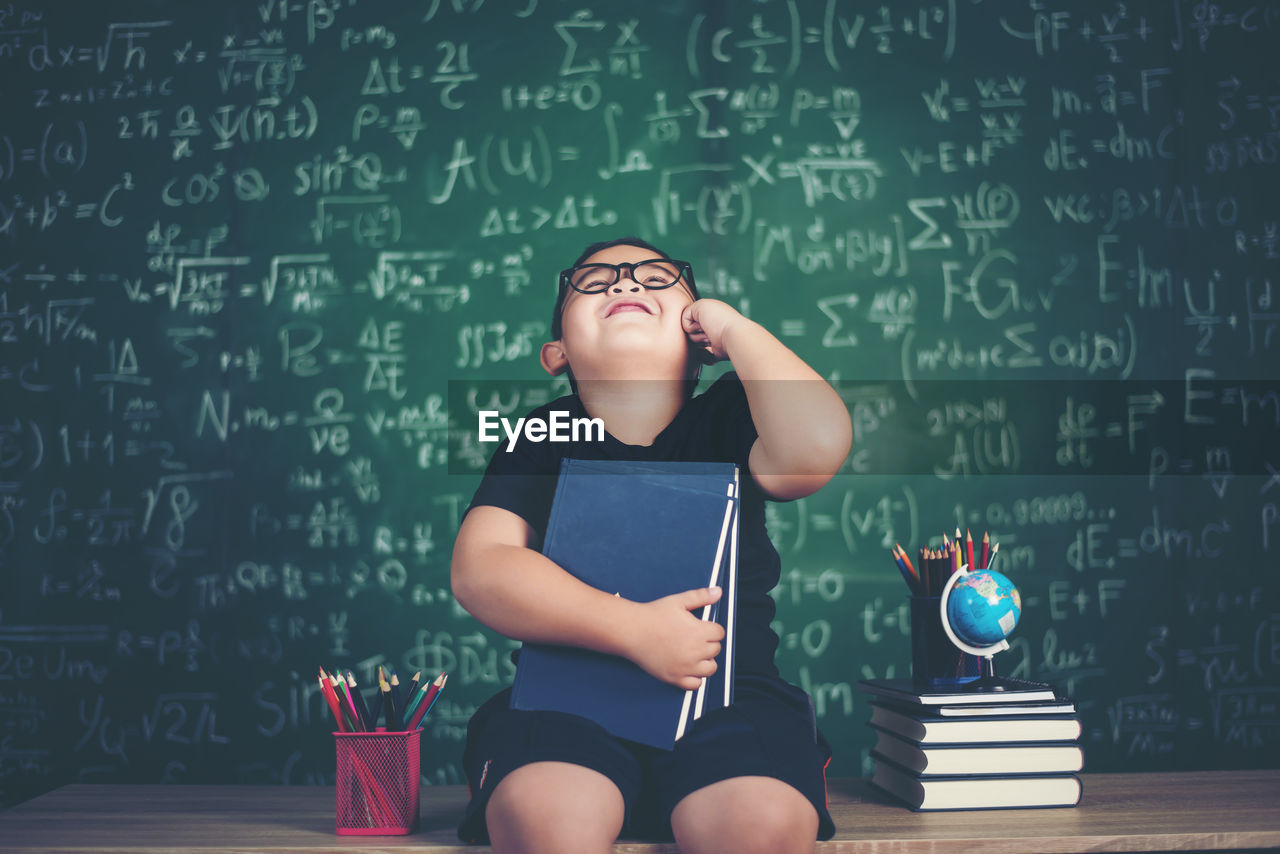 Cute boy holding books while sitting at table against blackboard