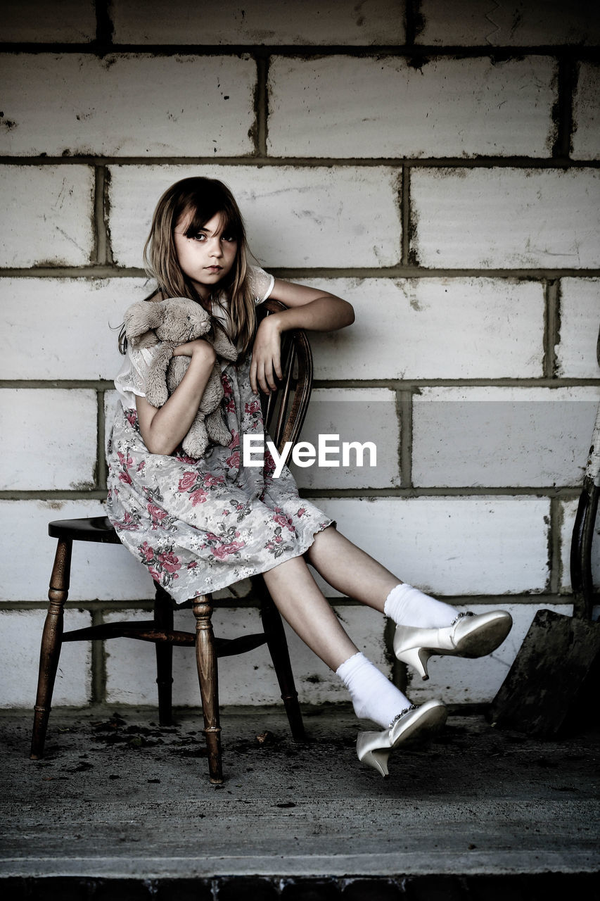 Portrait of girl holding stuffed toy and sitting on chair against wall