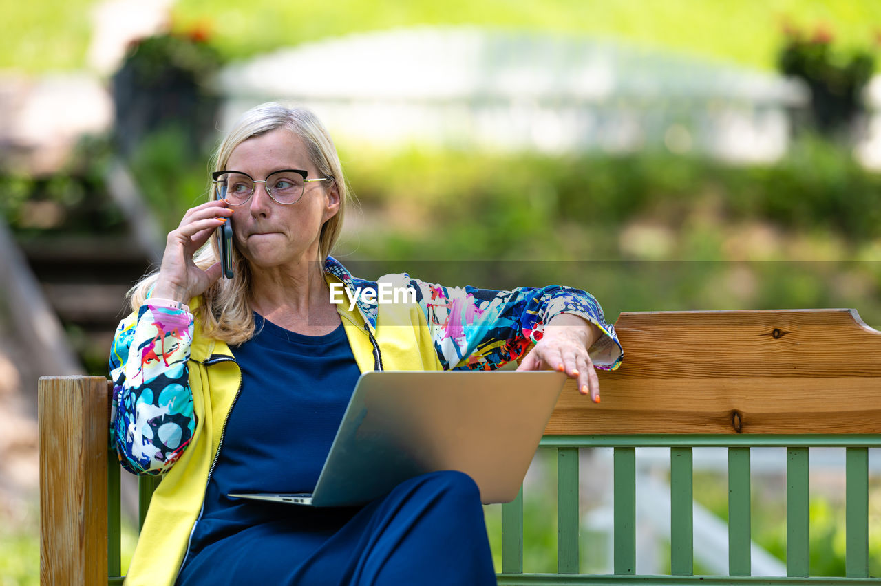 Senior woman at the garden working from home using laptop and speaking on the phone