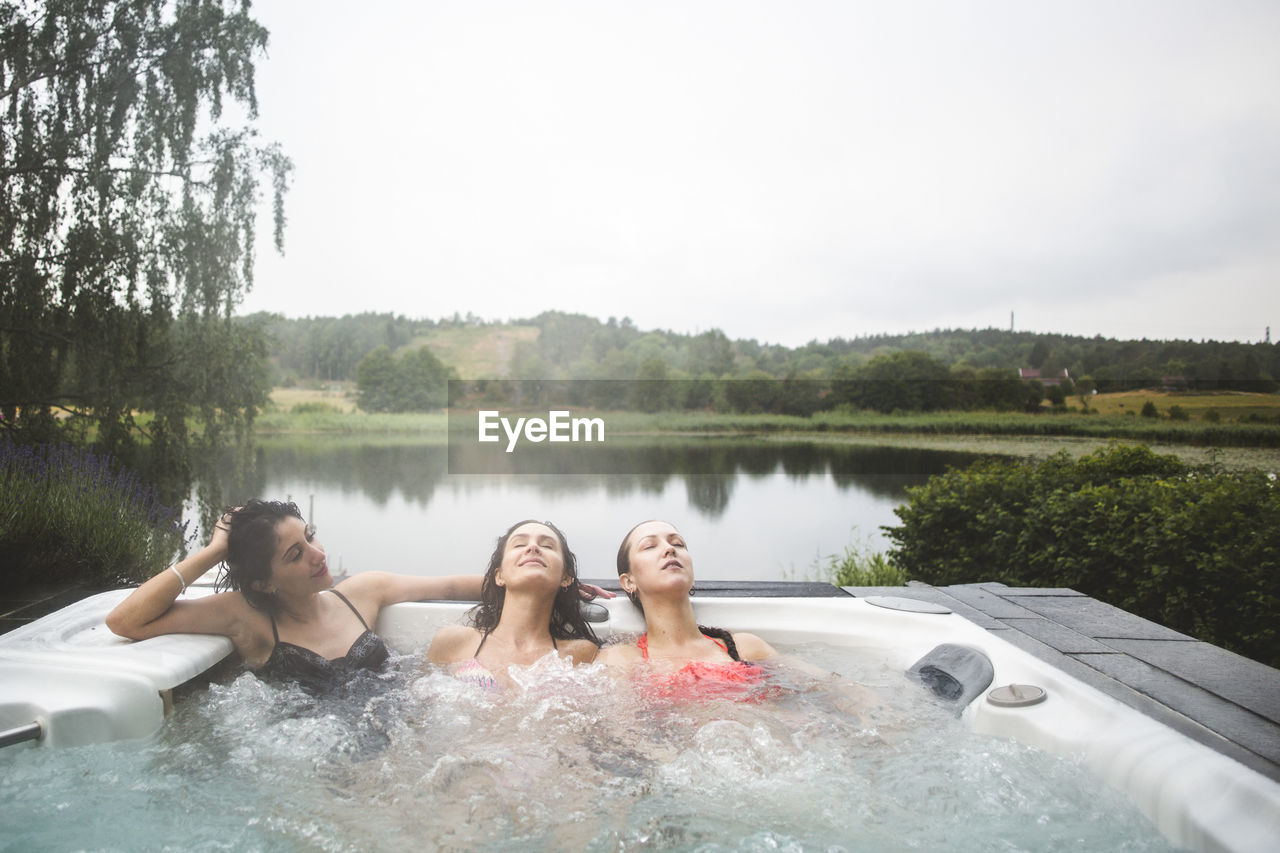 Female friends enjoying in hot tub with eyes closed against lake during weekend getaway