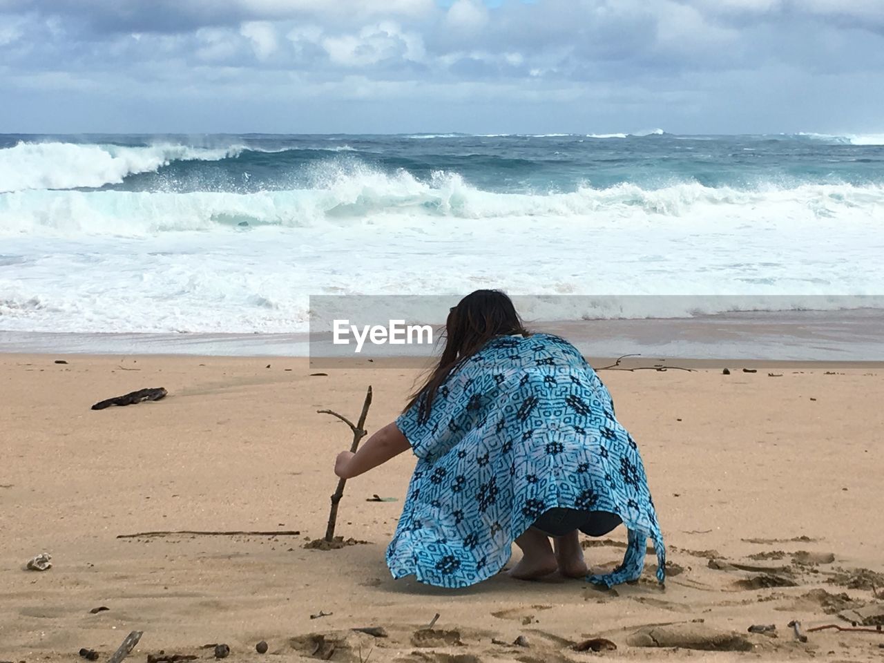 Rear view of woman crouching at beach against cloudy sky