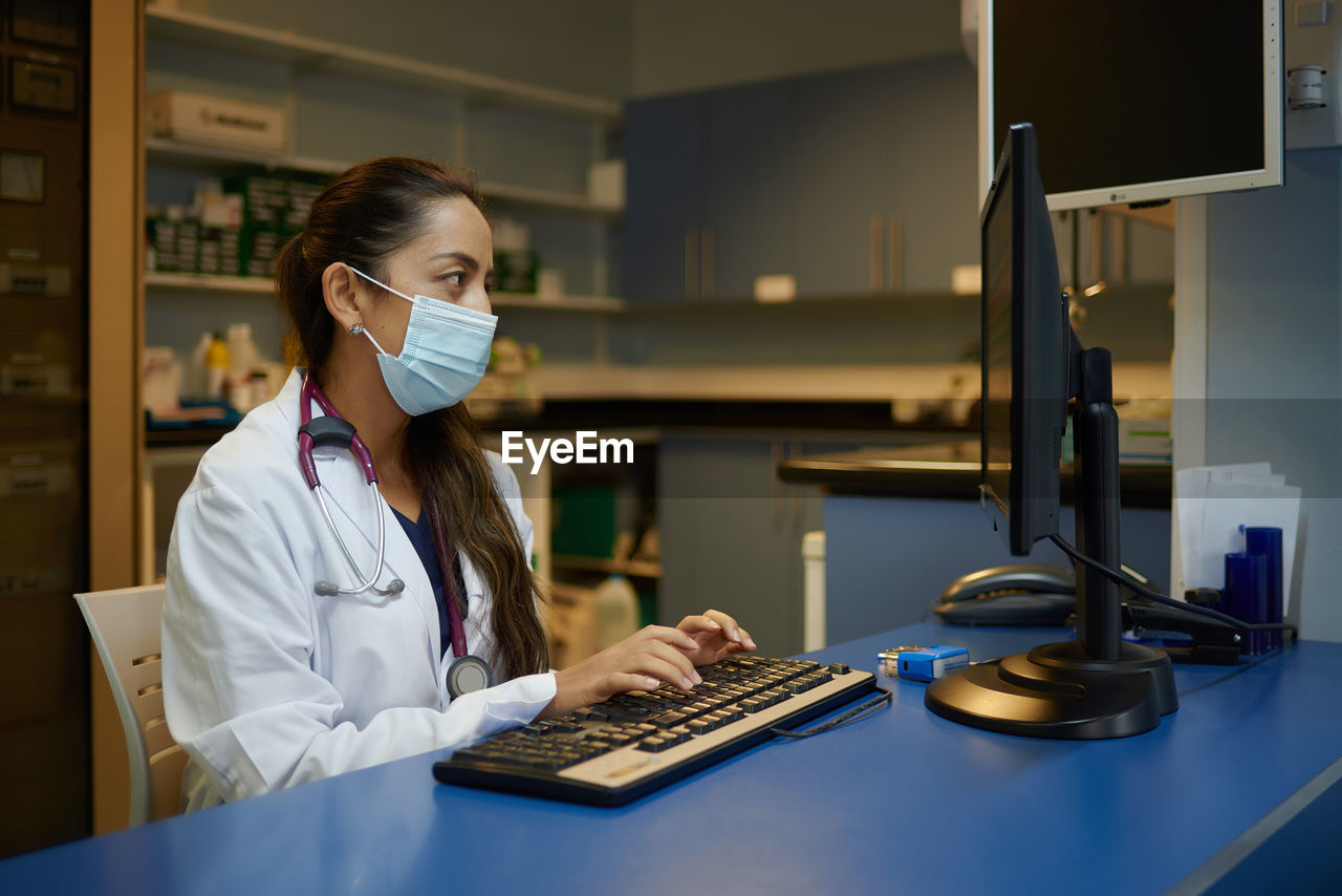 Side view of young female medic in white coat and protective mask typing on computer keyboard while working in cabinet of modern clinic