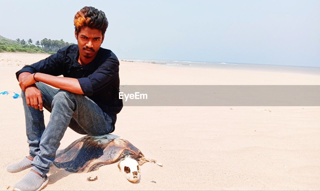 Young man sitting on dead turtle skeleton at beach against sky