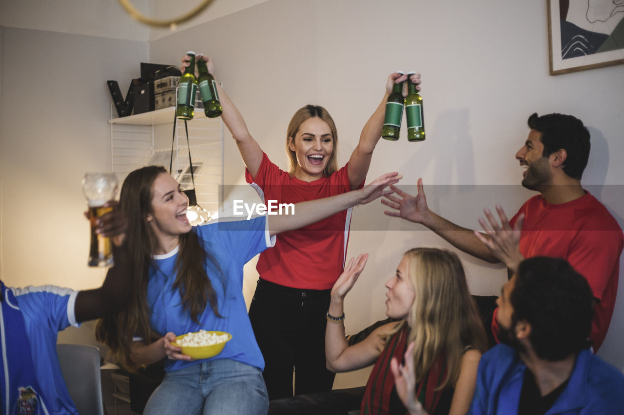 Cheerful woman holding beer bottles standing with friends in living room