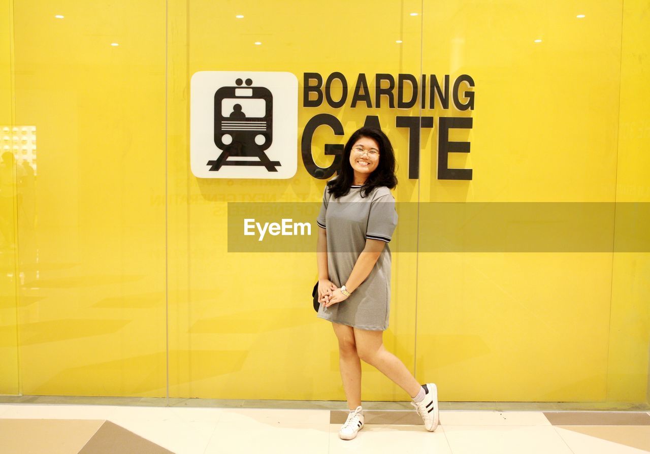 Portrait of smiling young woman standing against yellow wall with text at subway station