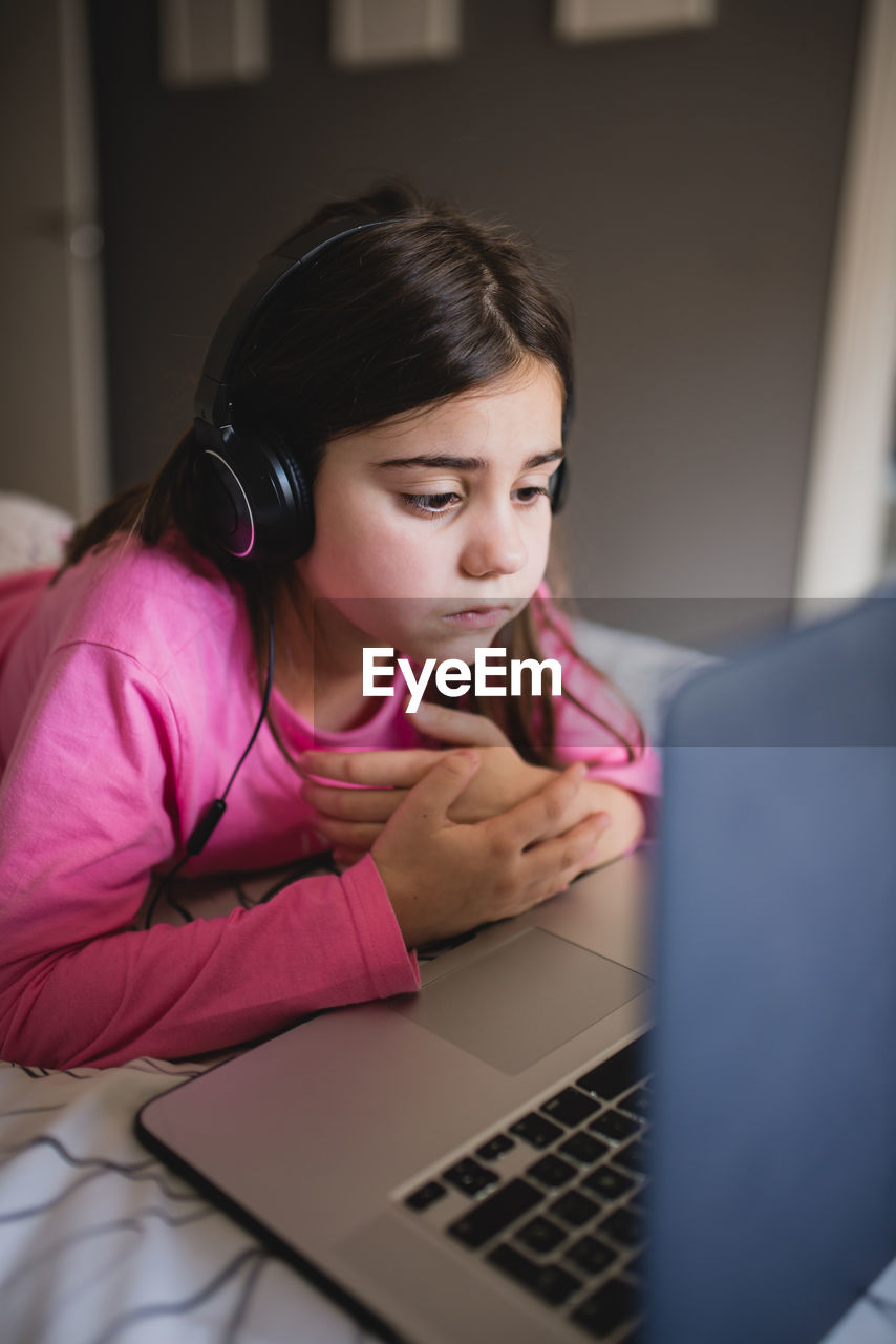 Focused little girl in headphones lying on bed and watching online video on laptop while spending free time at home