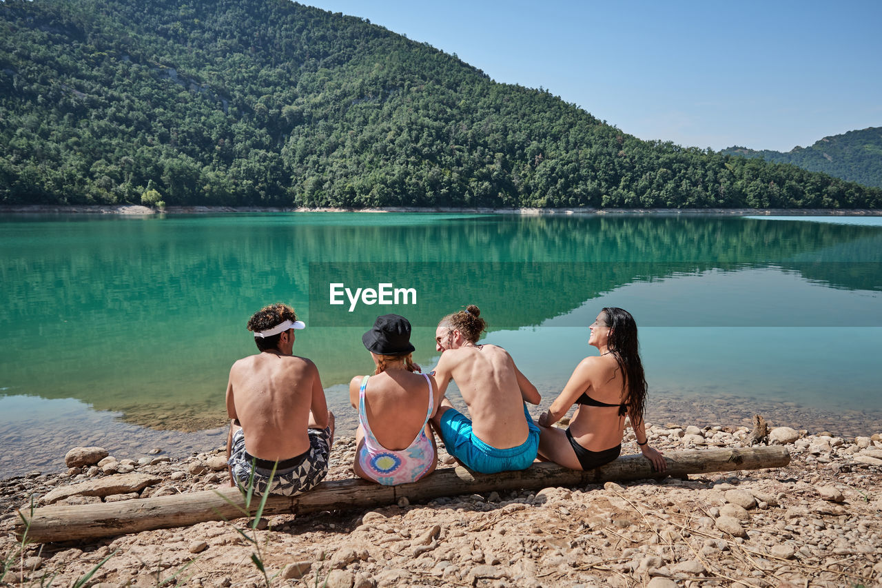 Back view of company of friendly people relaxing on tree log near lake and enjoying sunny day during vacation together