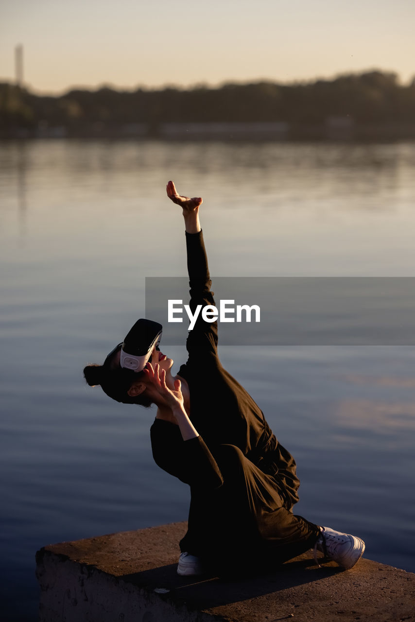 side view of woman with arms raised on beach against sky during sunset