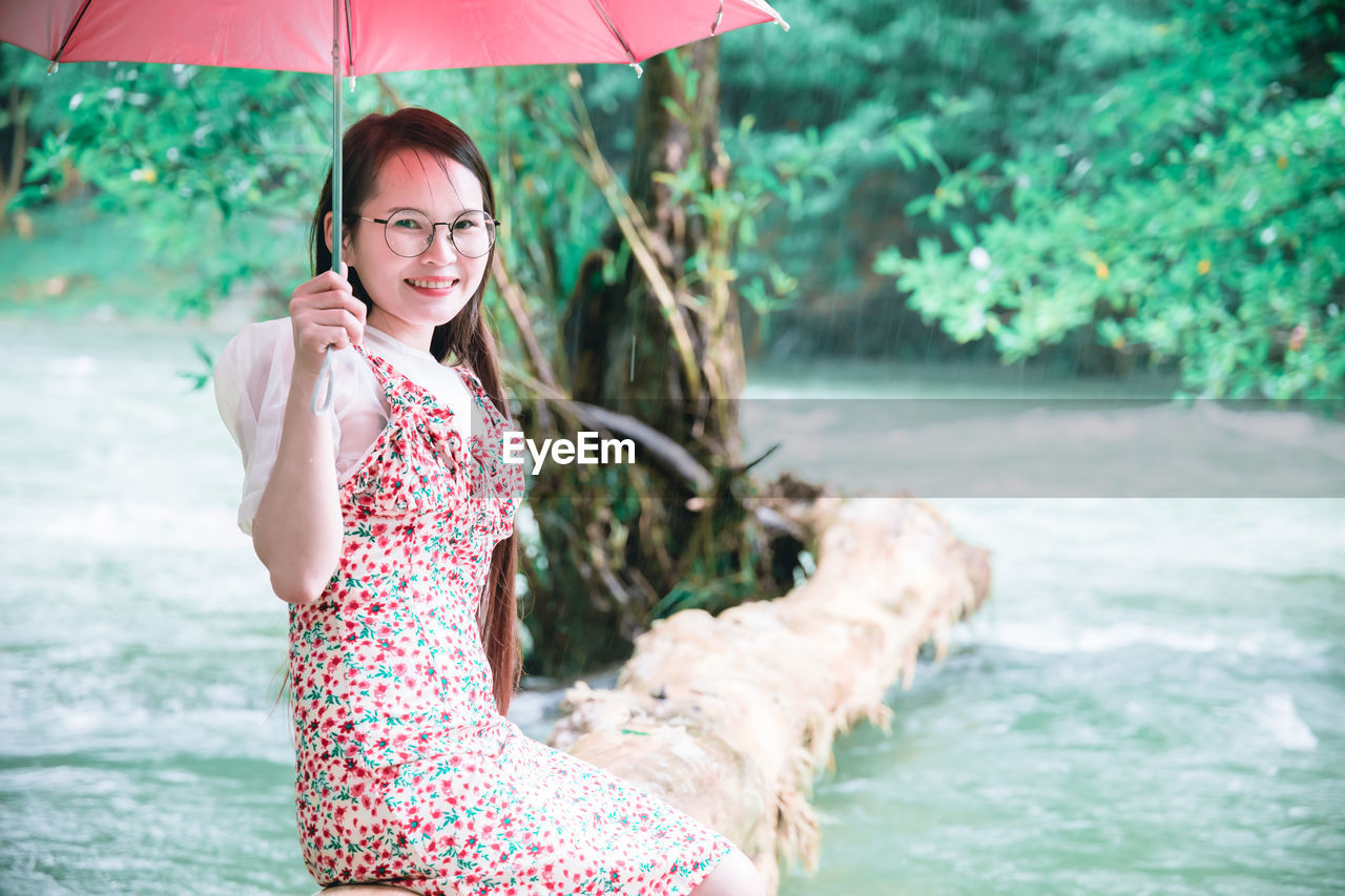 Portrait of smiling young woman holding umbrella sitting by river