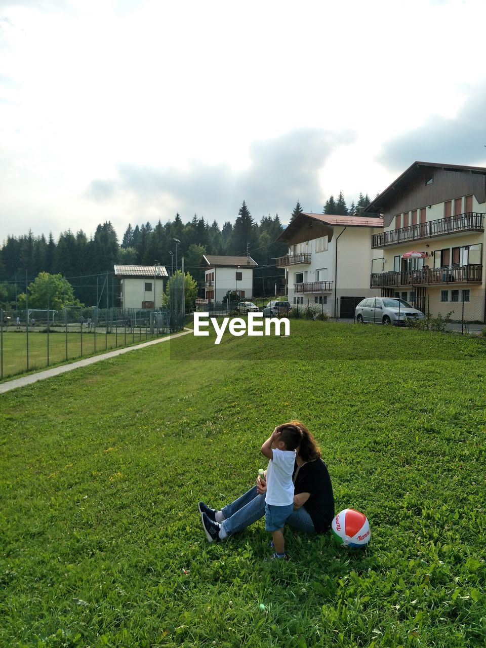 REAR VIEW OF COUPLE SITTING ON GRASSY FIELD AGAINST BUILDINGS