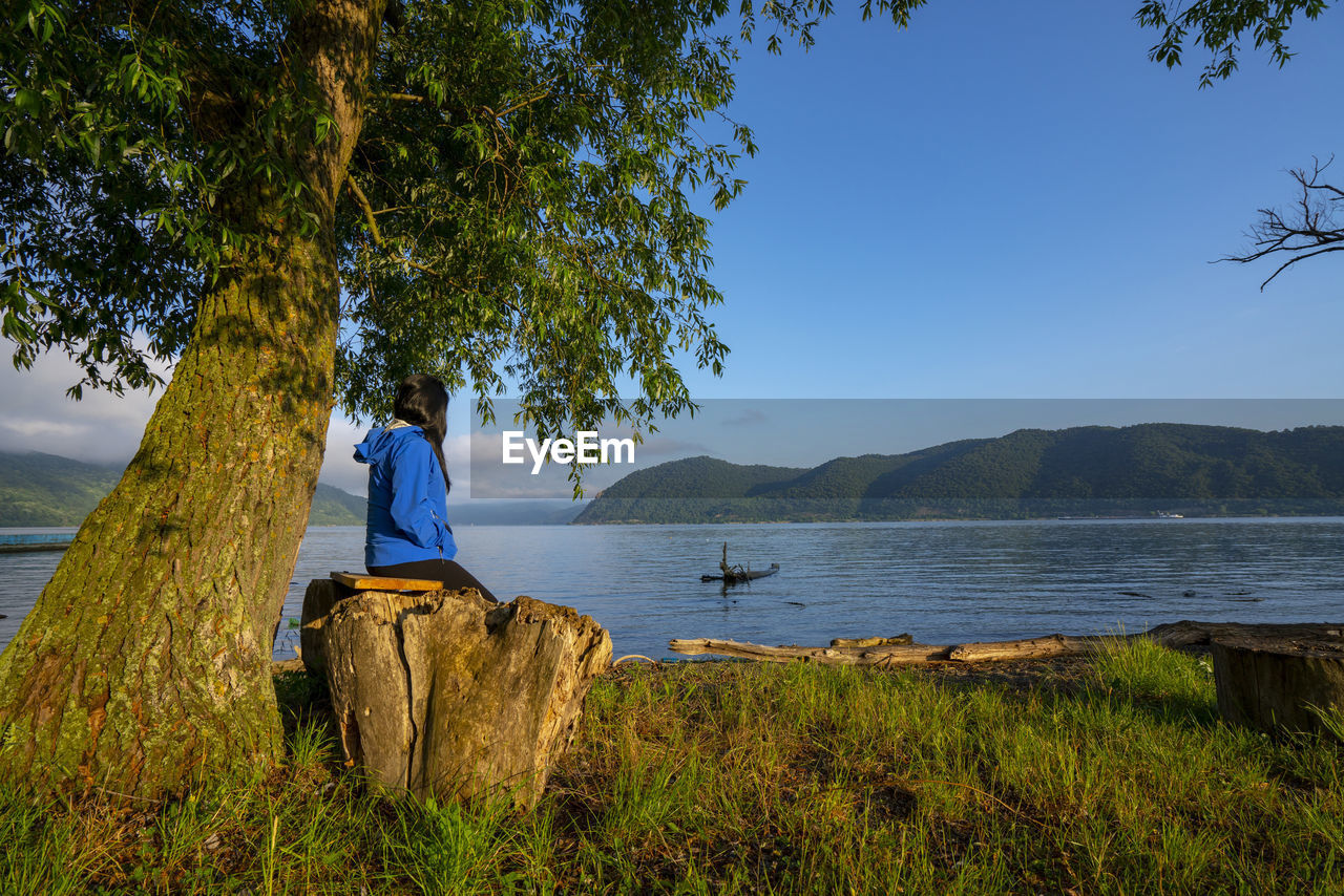 Side view of woman sitting by tree trunk looking at view of river against sky