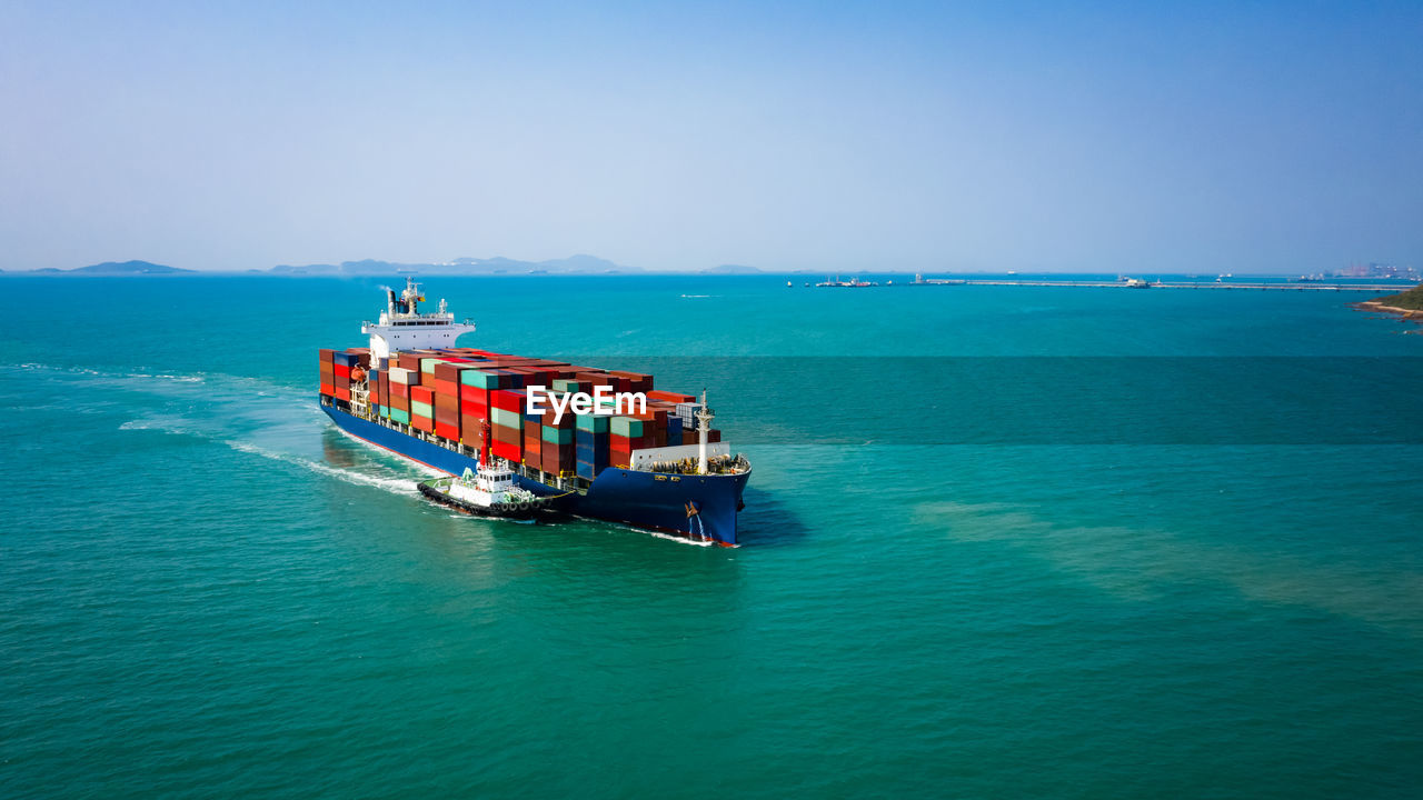 Container ship sailing in green sea and tugboat drag and blue sky background from drone shot