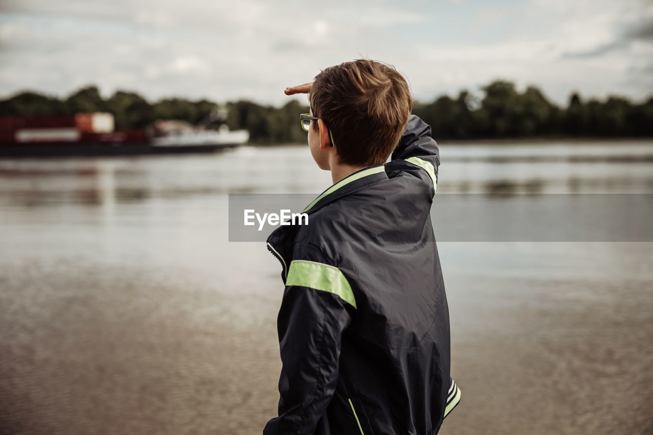 Young teen boy looking for container shipment on river with hand on head to protect eyes from sun