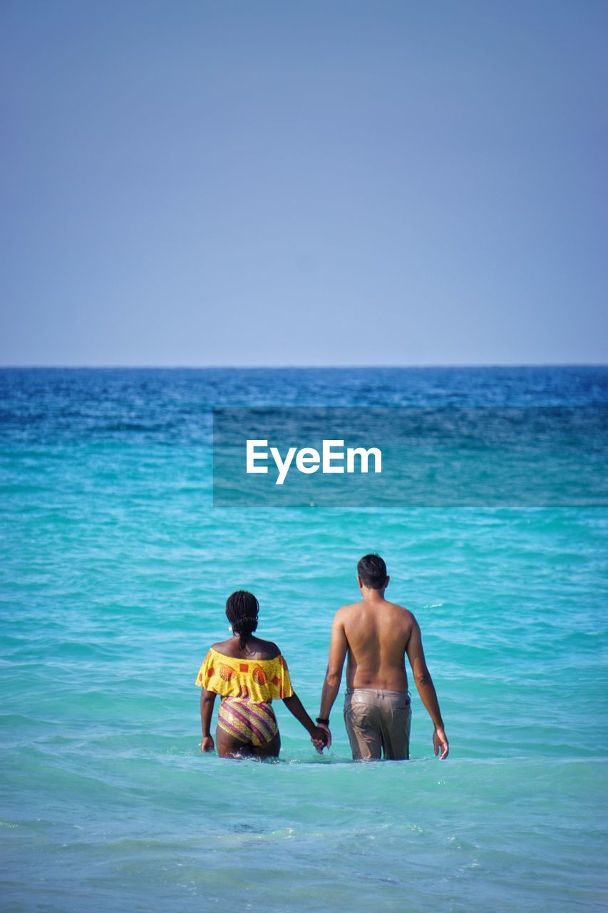 Shirtless man holding hand of woman while standing in sea against clear blue sky