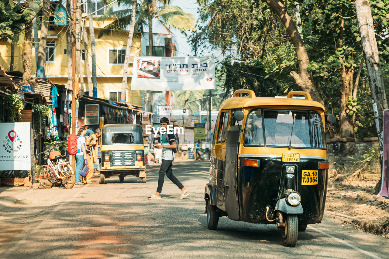 transportation, mode of transportation, city, street, rickshaw, transport, tree, architecture, land vehicle, jinrikisha, road, vehicle, car, motor vehicle, day, nature, plant, taxi, men, travel, building exterior, sunlight, city life, city street, adult, outdoors, built structure, public transportation, group of people, urban area, bus