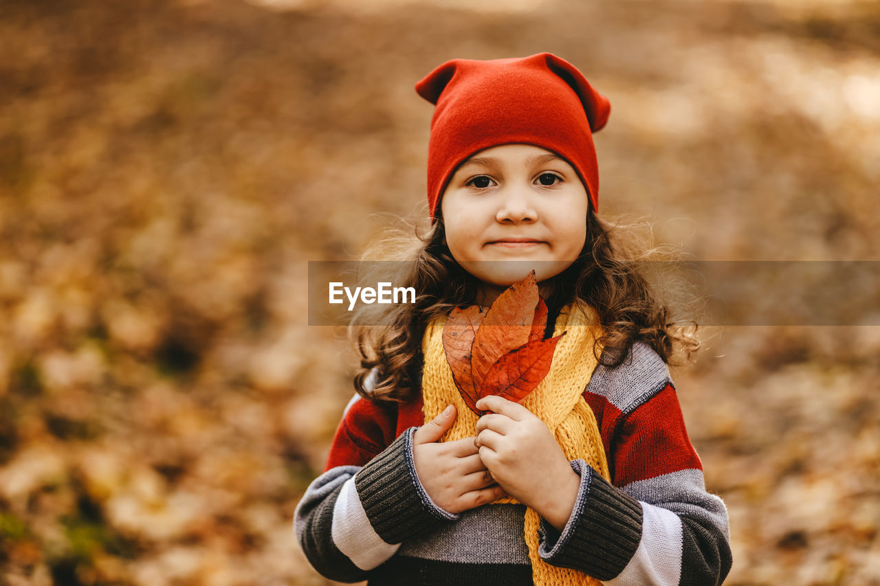 Portrait of a little girl a child in a warm hat walking holding an autumn leaf in the fall forest