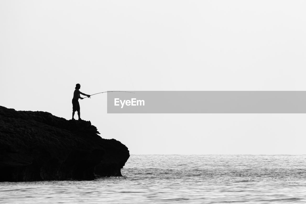 Man standing on rock fishing against clear sky