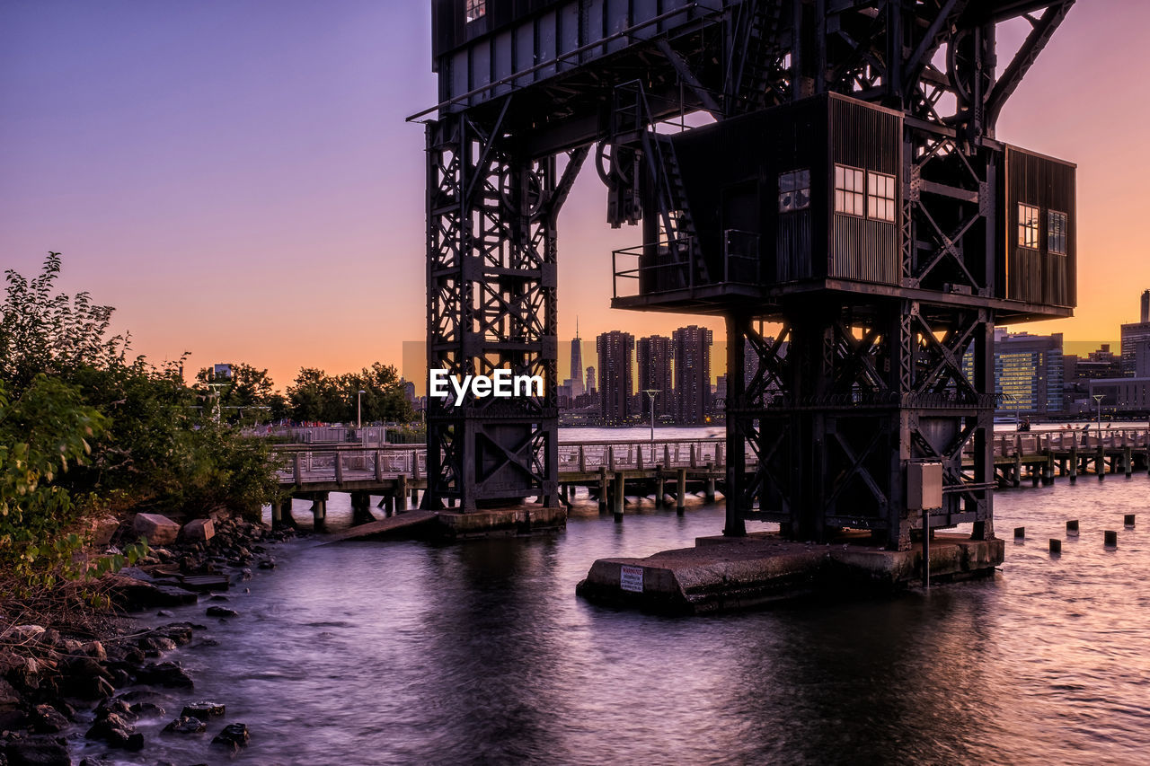 BRIDGE OVER RIVER IN CITY AGAINST SKY AT SUNSET