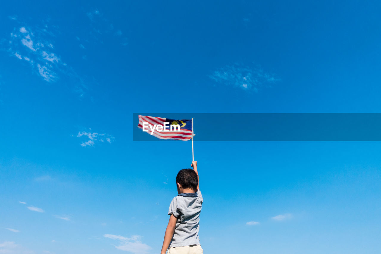 Low angle view of boy holding flag while standing against blue sky