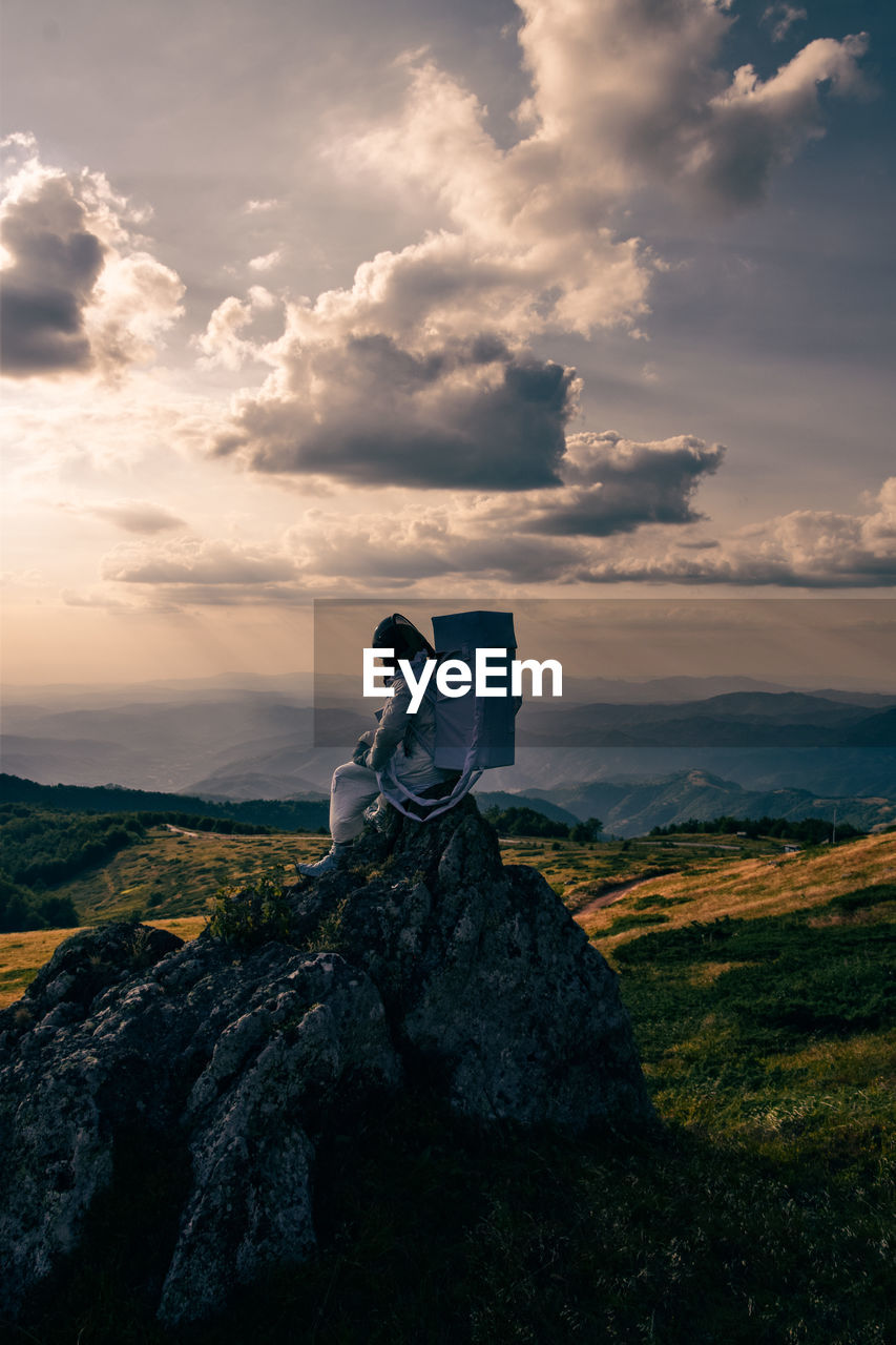 Rear view of man sitting on rock against sky during sunset