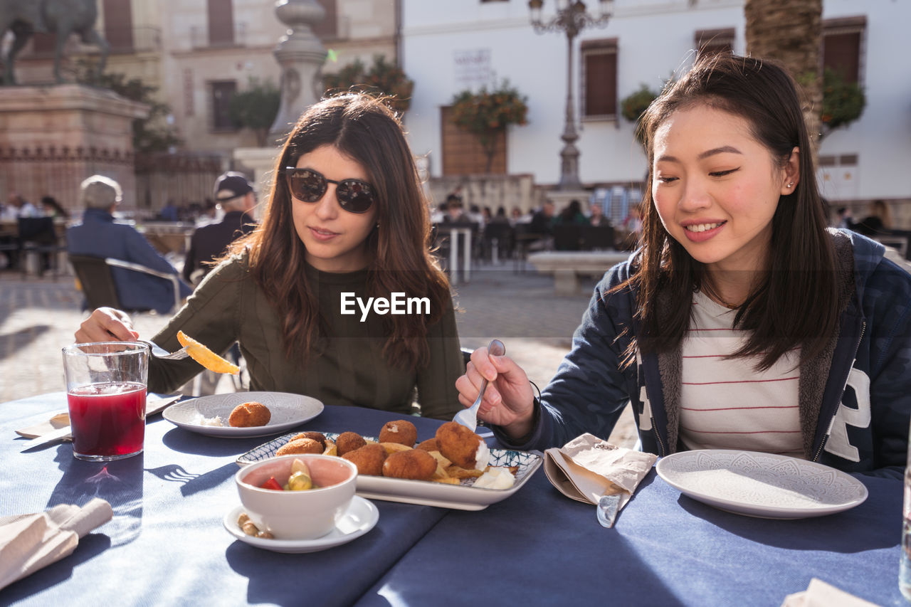 Content asian woman with female friend eating fried nuggets while having lunch together at table in outdoor cafeteria on sunny day