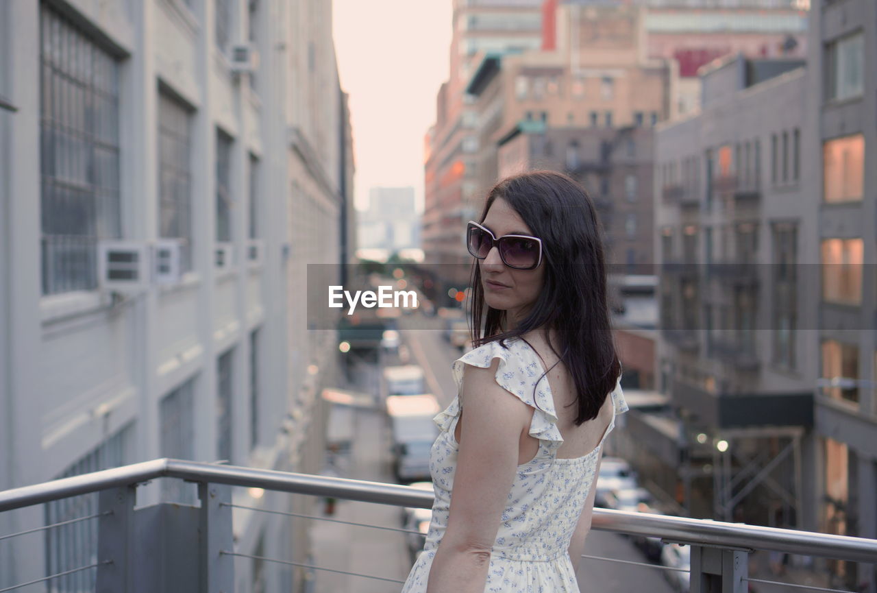 Portrait of woman wearing sunglasses while standing at balcony in city during sunset