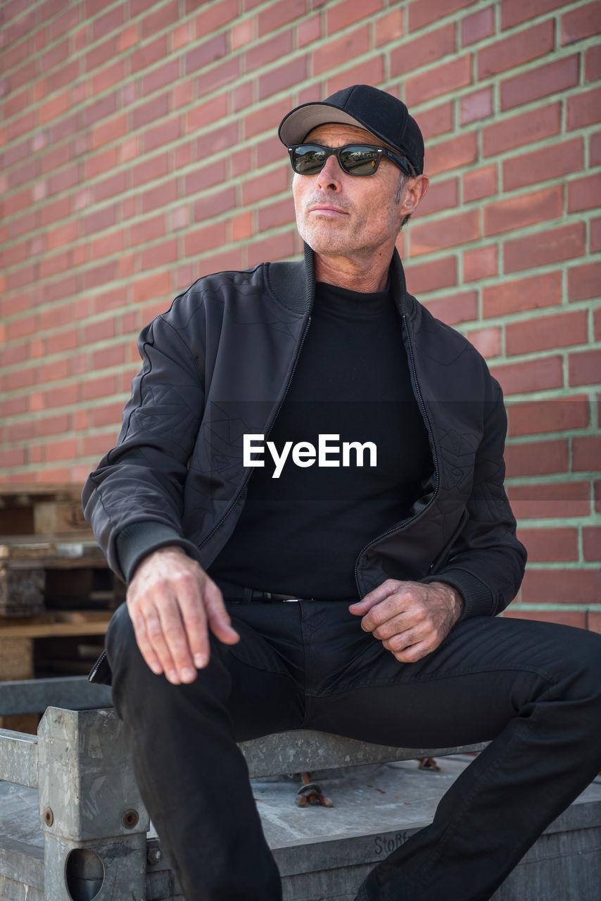 Man wearing black jacket and sunglasses while sitting against brick wall