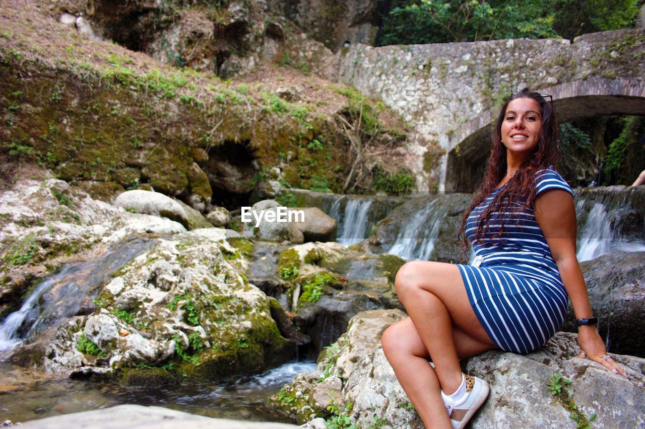 SMILING YOUNG WOMAN SITTING ON ROCK AGAINST PLANTS