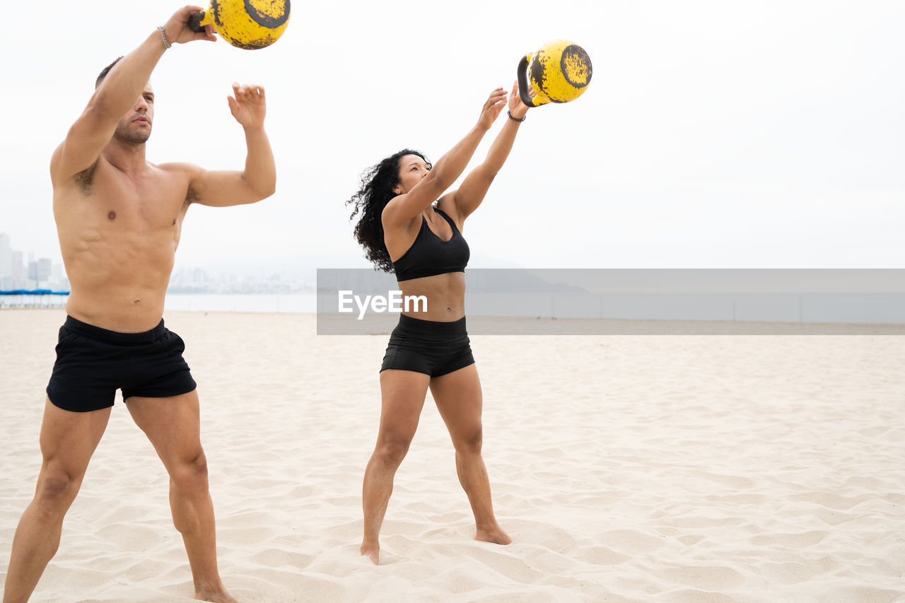 Multiracial fit sportsman and sportswoman doing kettlebell swing exercise while training at seaside in summer