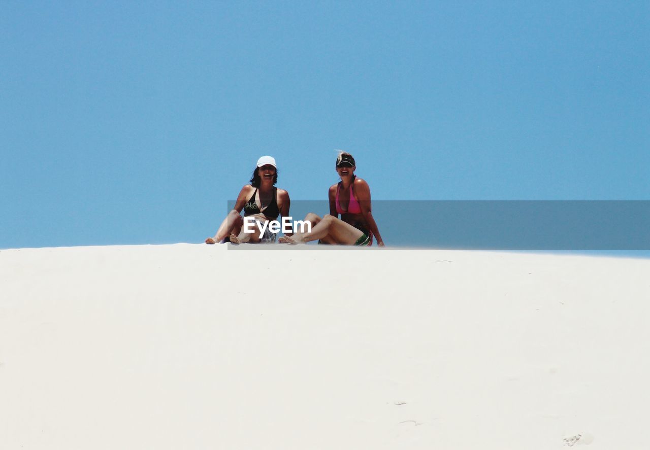 Friends sitting on sand dune against clear blue sky during sunny day